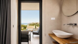Un baño moderno con un lavabo de pedestal grande, un espejo redondo y vistas al mar a través de una puerta abierta.