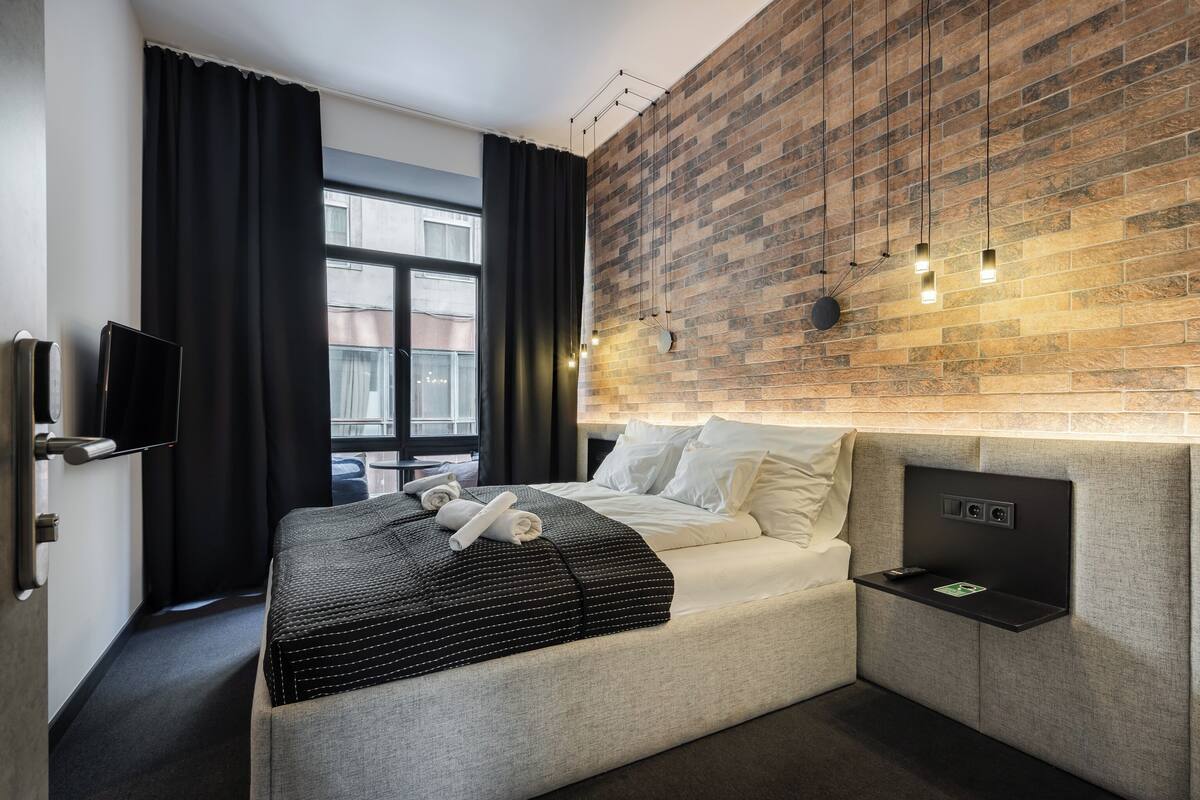 A modern hotel room with a brick-patterned wall, a flat-screen TV, a bedside table with a remote control, and a neatly made bed with a striped blanket.