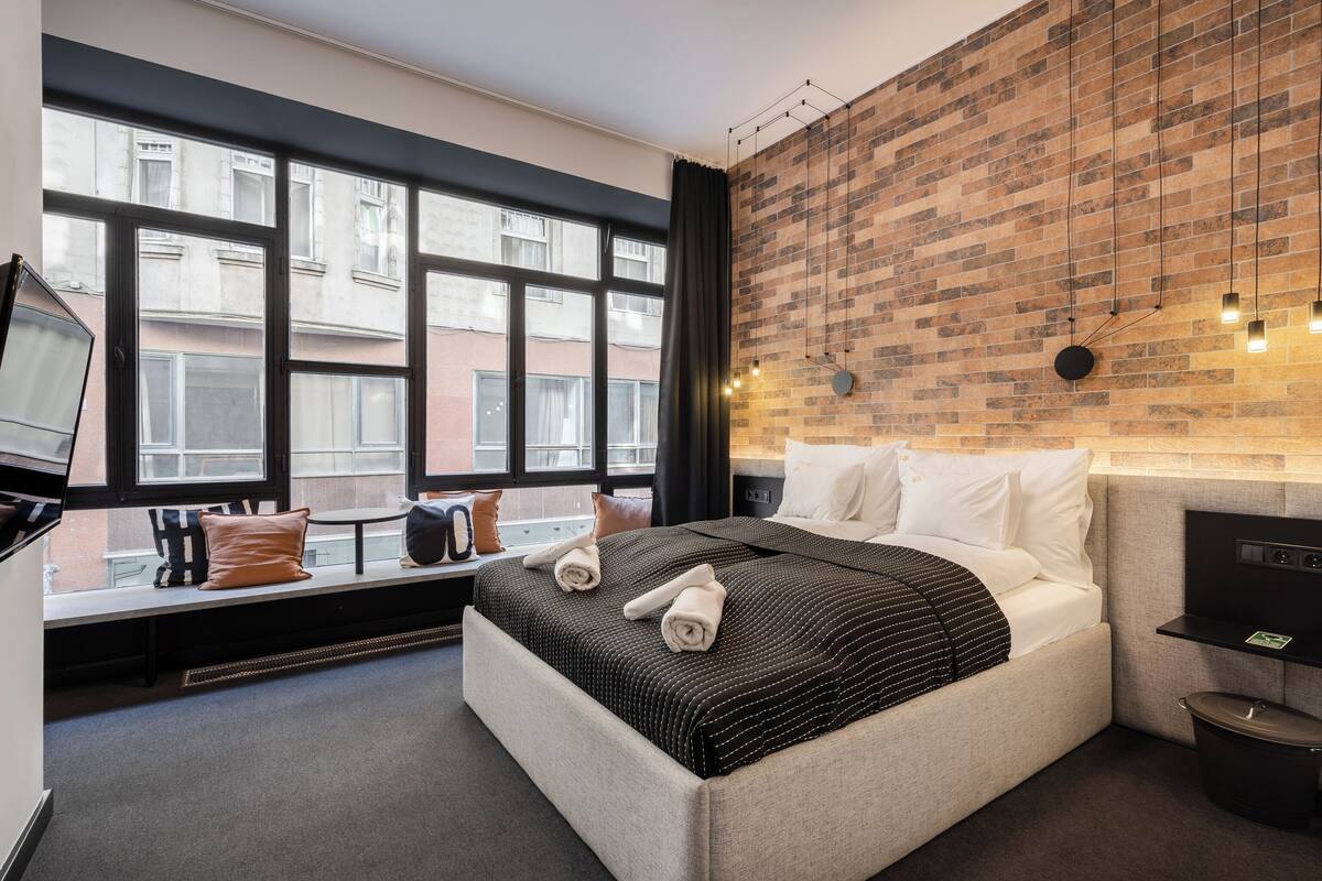 A modern hotel room with a large bed, a bench by the window, and a brick wall.