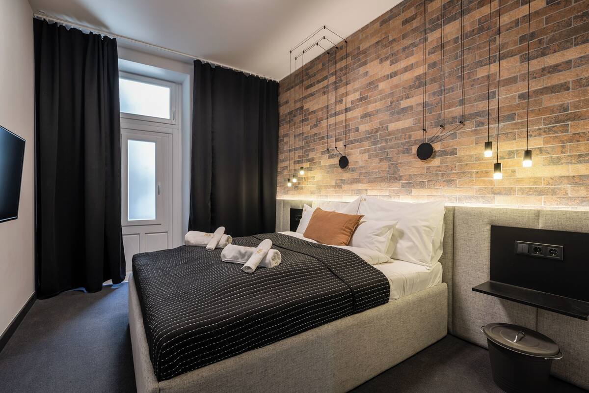 A modern hotel room with a brick-patterned wall, a bed with a black and white striped bedspread, a flat-screen TV, and a small kitchen area with a stove.