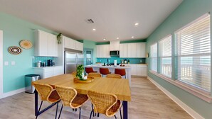 A modern kitchen with white cabinets, a stainless steel refrigerator, a microwave, and a dining area with a wooden table and wicker chairs. - Tiffany House: 4  BR, 3.5  BA House in Crystal Beach, Sleeps 16 (Bolivar Peninsula)