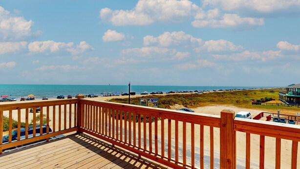 A wooden deck with a view of the beach, vehicles, and a distant building. - Tiffany House: 4  BR, 3.5  BA House in Crystal Beach, Sleeps 16 (Bolivar Peninsula)