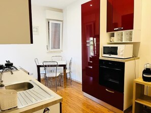 A compact kitchen with a sink, stove, and microwave. There is a dining table with chairs and a window with blinds. - LUDOVICA CASA VACANZE (Palermo)