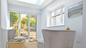 A modern living room with a grey armchair, a beige ottoman, and a glass door leading to an outdoor patio. - Grace Cottage - Emsworth -  a cottage that sleeps 6 guests  in 3 bedrooms (Emsworth)