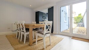 A dining area with a wooden table and white chairs, a kitchen in the background, and a large sliding glass door leading to an outdoor area. - Grace Cottage - Emsworth -  a cottage that sleeps 6 guests  in 3 bedrooms (Emsworth)