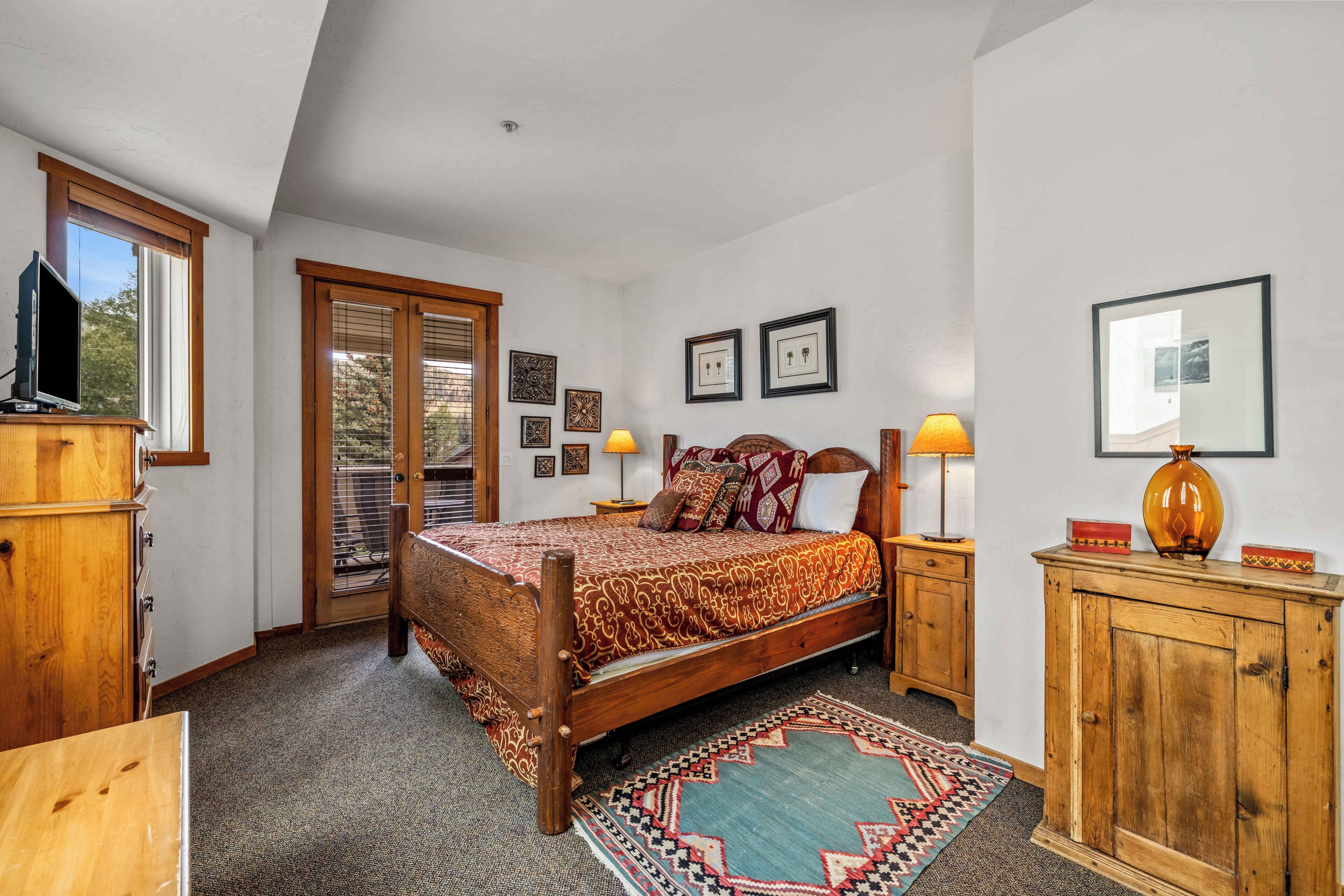 A bedroom with a wooden bed, a nightstand with a lamp, a dresser, and a window with blinds.