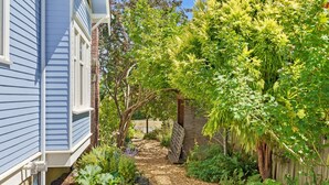 Un vialetto immerso nel verde che conduce a una struttura in legno, circondato da vegetazione lussureggiante e un edificio blu.
