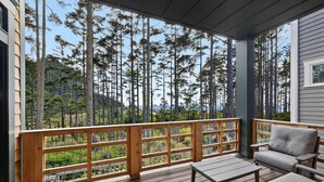 A wooden deck with a view of tall trees, a gray sofa, and a table. - Seabrook Seaclusion | Pet Friendly, Ocean View 4BR | Sleeps 10, Elevator (Pacific Beach)
