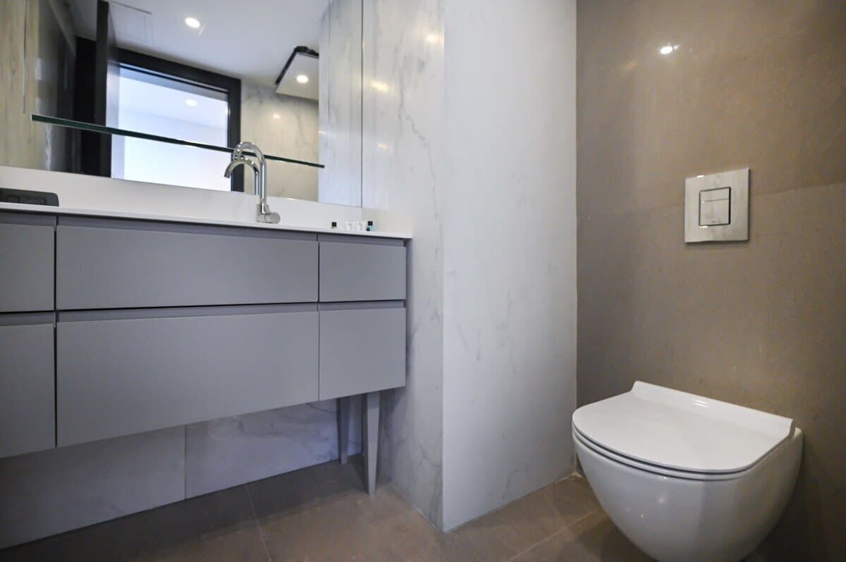 A modern bathroom with a white vanity, a mirror, a toilet, and a light switch on the wall.