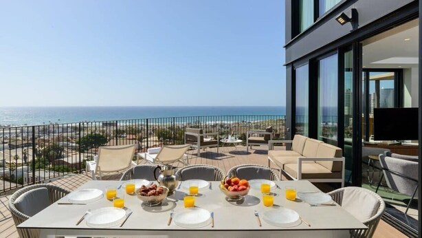 Une table à manger dressée avec des assiettes, des couverts et des corbeilles de fruits, donnant sur une plage et l’océan.