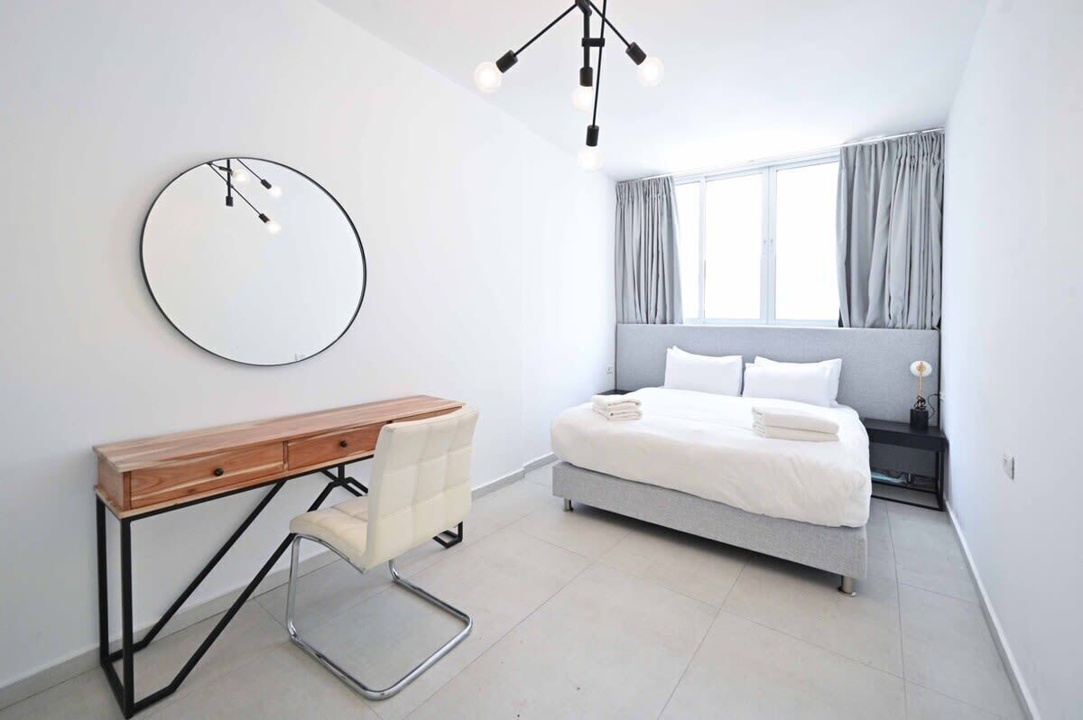 A modern bedroom with a bed, bedside table, chair, and a round mirror on the wall.