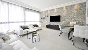 Un salon moderne avec un mur d’accent en pierre, un téléviseur à écran plat, un canapé blanc, une table à manger avec des chaises et une table basse en verre.