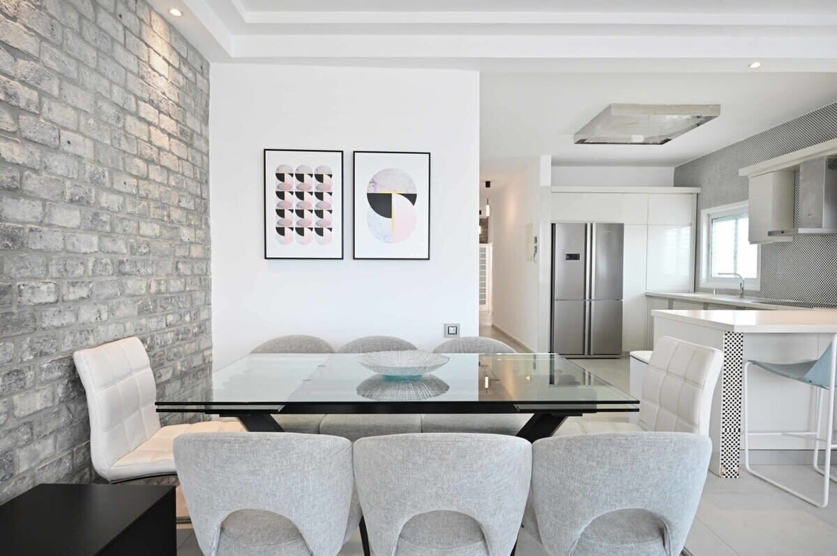 A modern dining area with a glass table, grey upholstered chairs, and a brick wall.