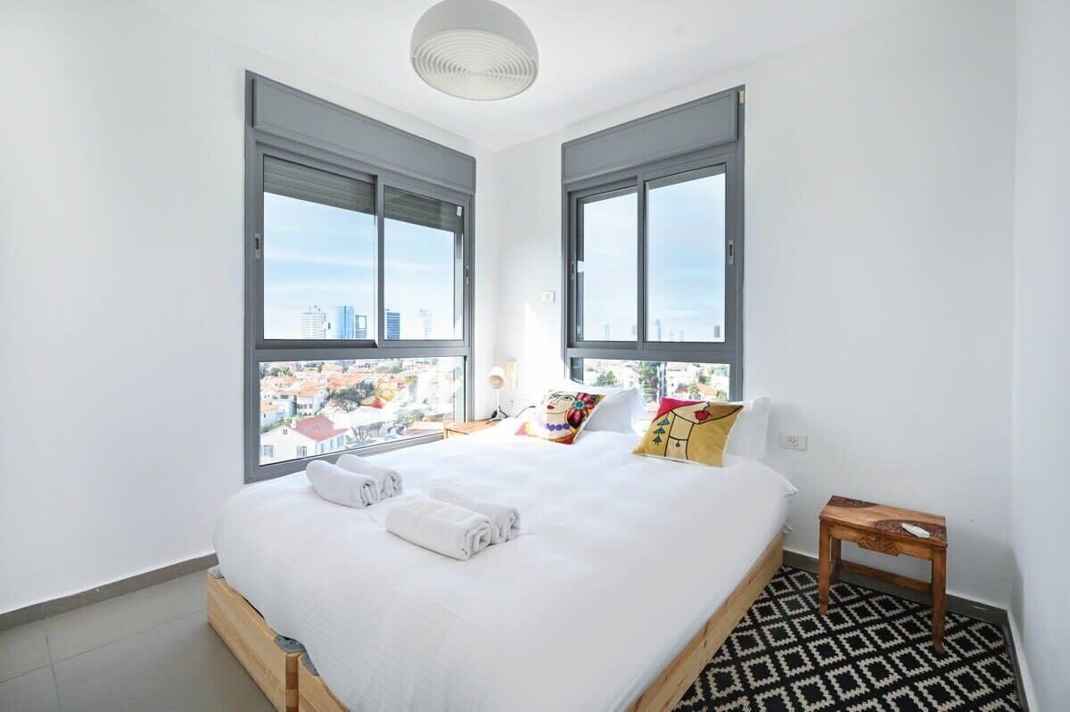 A modern hotel room with a large bed, two windows with city views, a wooden bedside table, and a patterned rug.