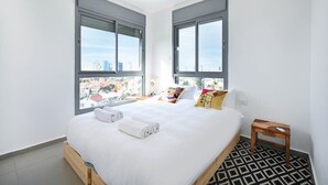 Une chambre d’hôtel moderne avec un grand lit, deux fenêtres donnant sur la ville, une table de chevet en bois et un tapis à motifs.
