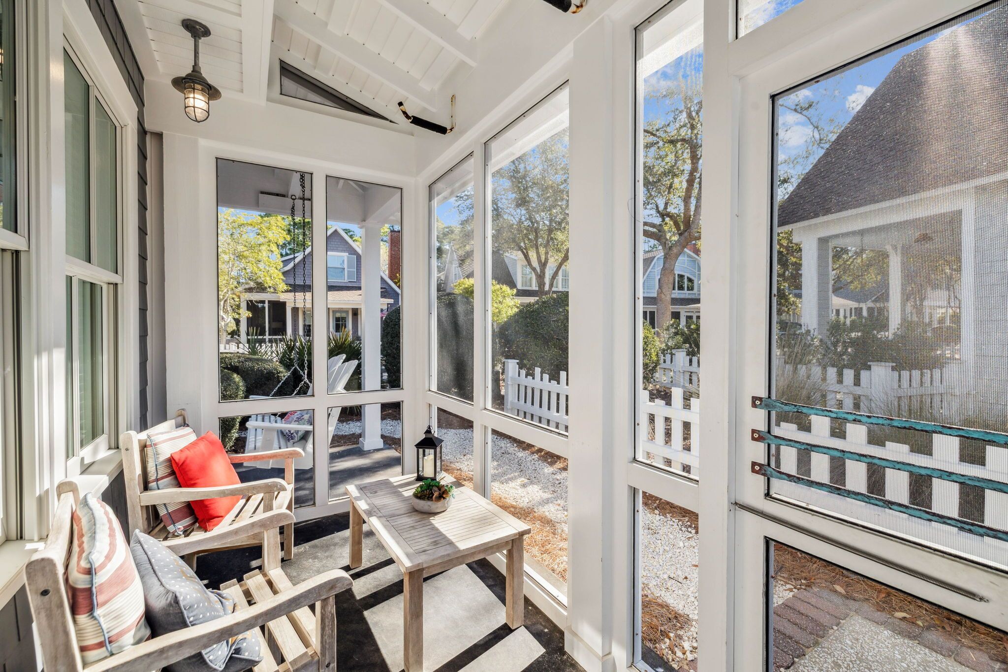 A sunroom with large windows, a wooden bench, a table with a lantern, and a view of a white picket fence and houses.