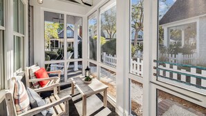 Uma sala de sol com grandes janelas, um banco de madeira, uma mesa com um candeeiro e vista para uma cerca branca e casas.