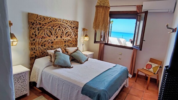 Ein Schlafzimmer mit einem großen Bett, einem dekorativen Kopfteil, einem Nachttisch, einem Sessel und einem Fenster mit Blick auf das Meer.