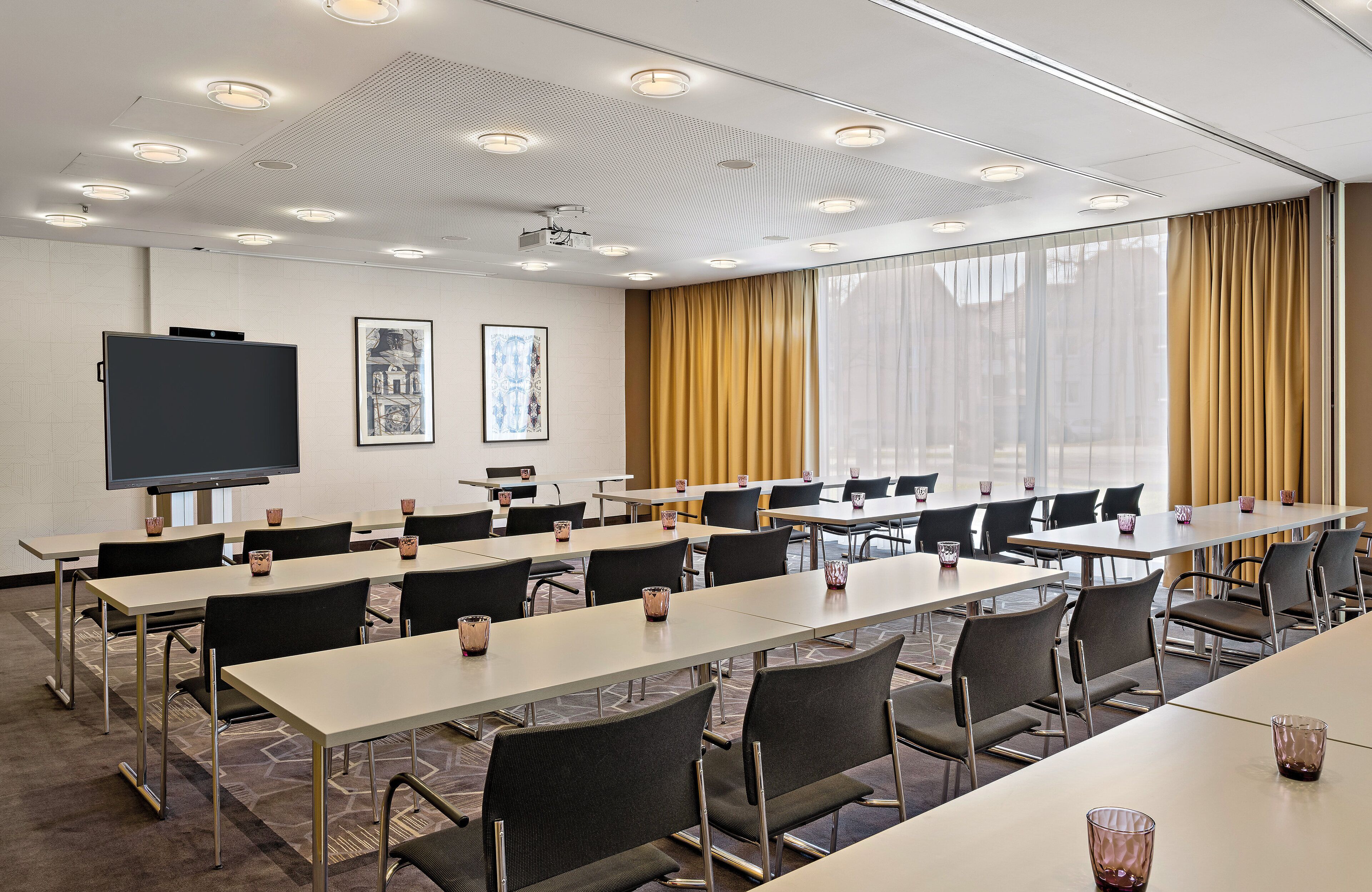 A conference room with multiple tables and chairs, a projector screen, and large windows with curtains.