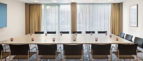 Sala de conferências com uma mesa comprida, cadeiras e uma grande janela com cortinas.