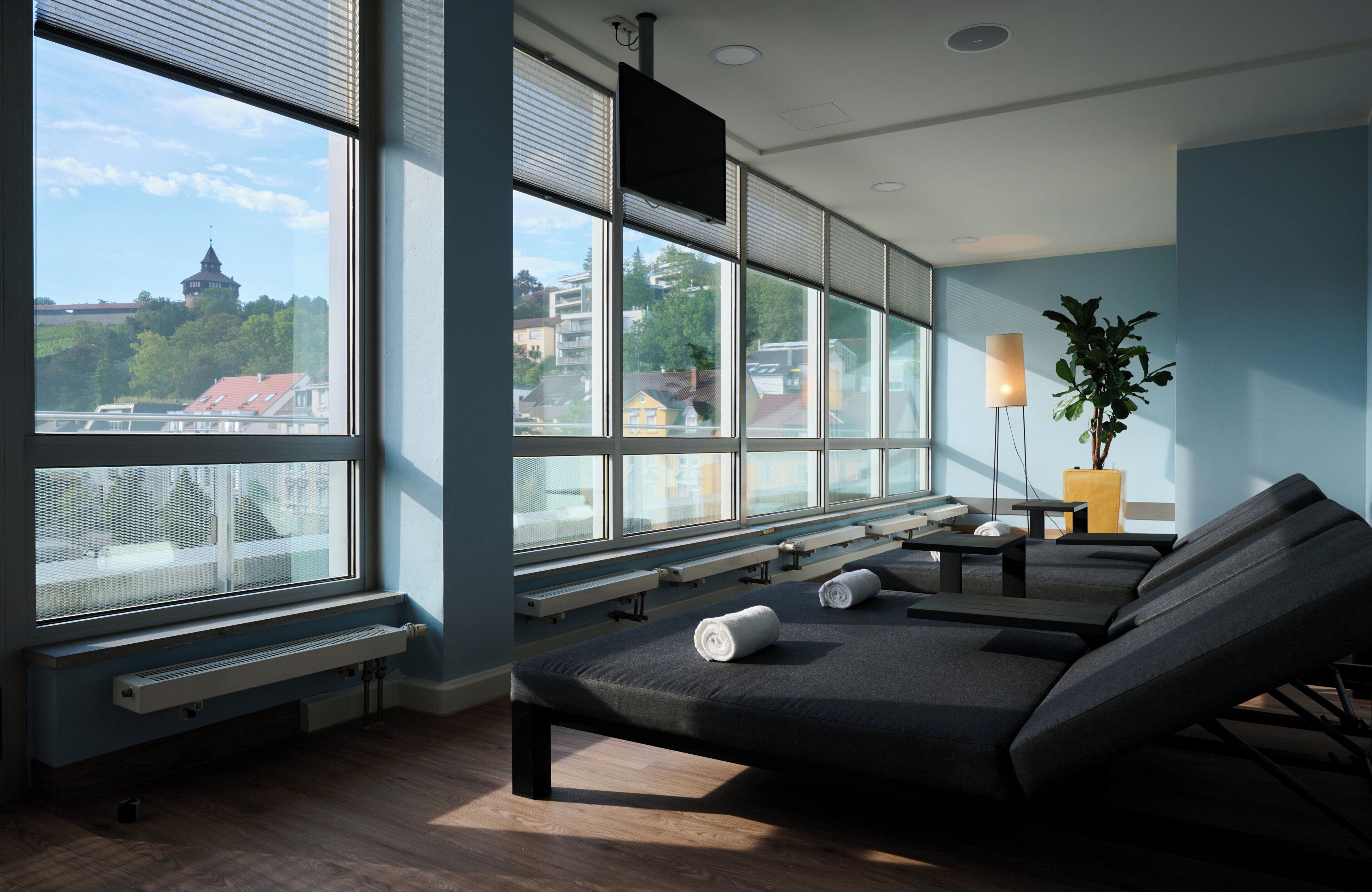 A modern spa room with large windows, a massage table, benches, and a potted plant.