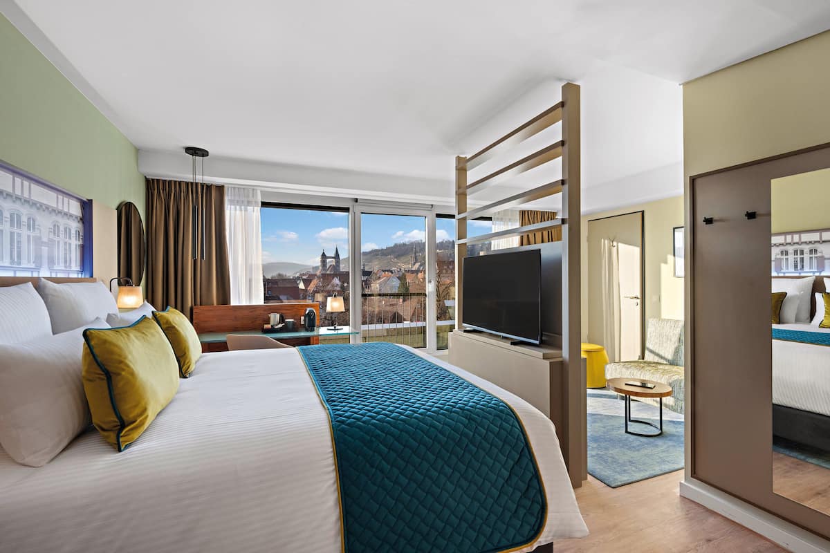 A hotel room with a large bed, a TV, a balcony with a city view, and a wooden ladder.