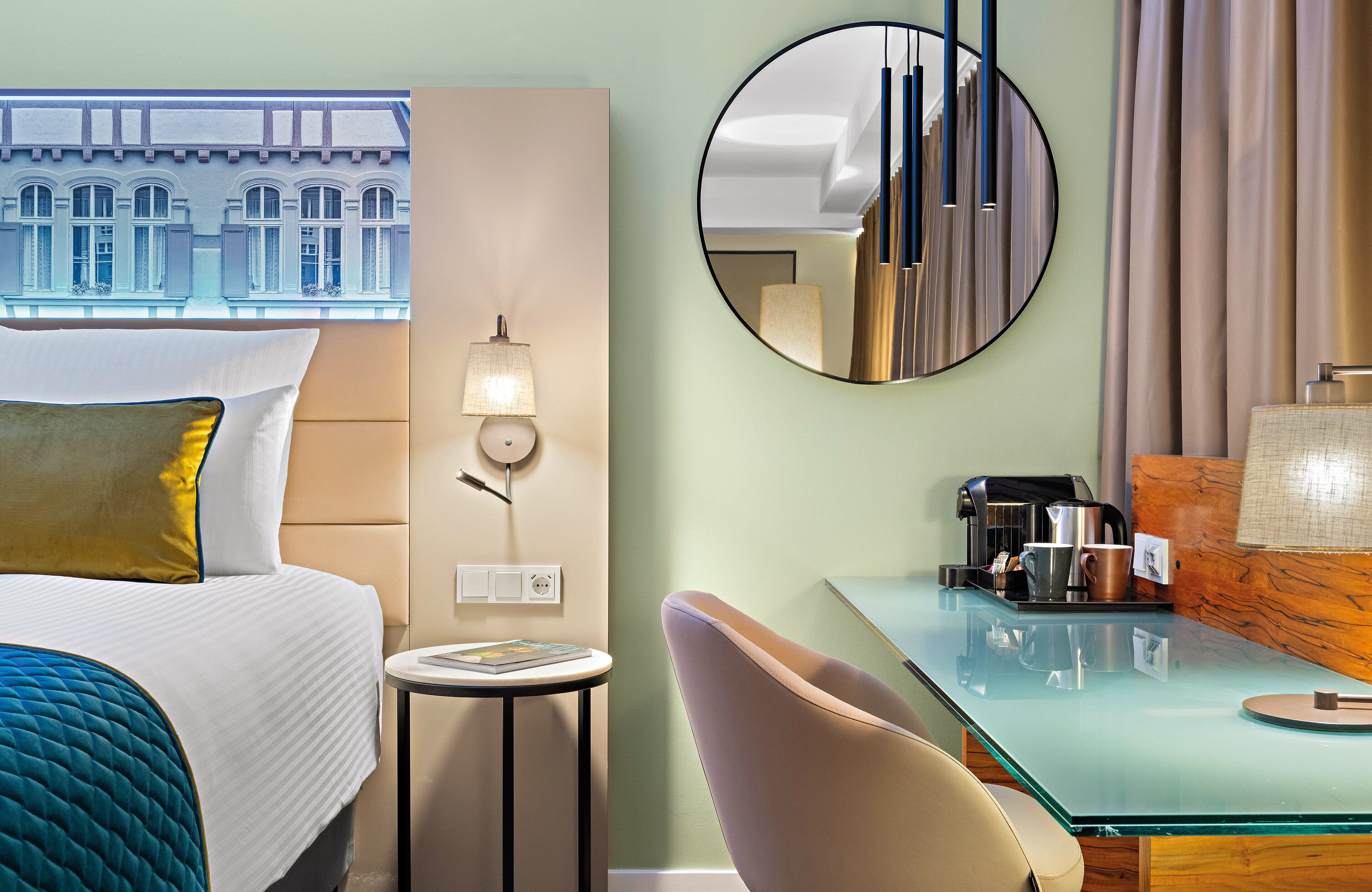Ett hotellrum med en säng, en rund spegel, ett skrivbord med en kaffebryggare och utsikt över en byggnad genom ett fönster.