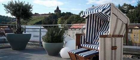 A balcony with a wicker chair, potted plants, and a view of a hill with a tower.