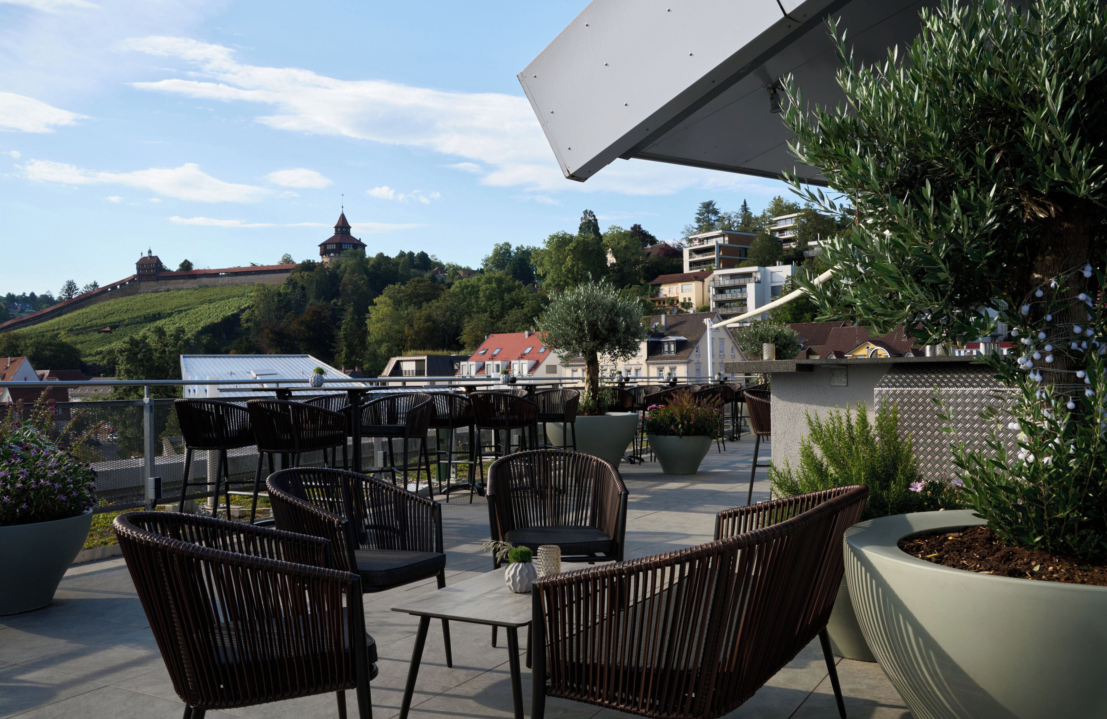 Un espace extérieur aménagé avec des chaises et des tables en osier, entouré de plantes en pot et offrant une vue sur une colline où se dressent des bâtiments en arrière-plan.