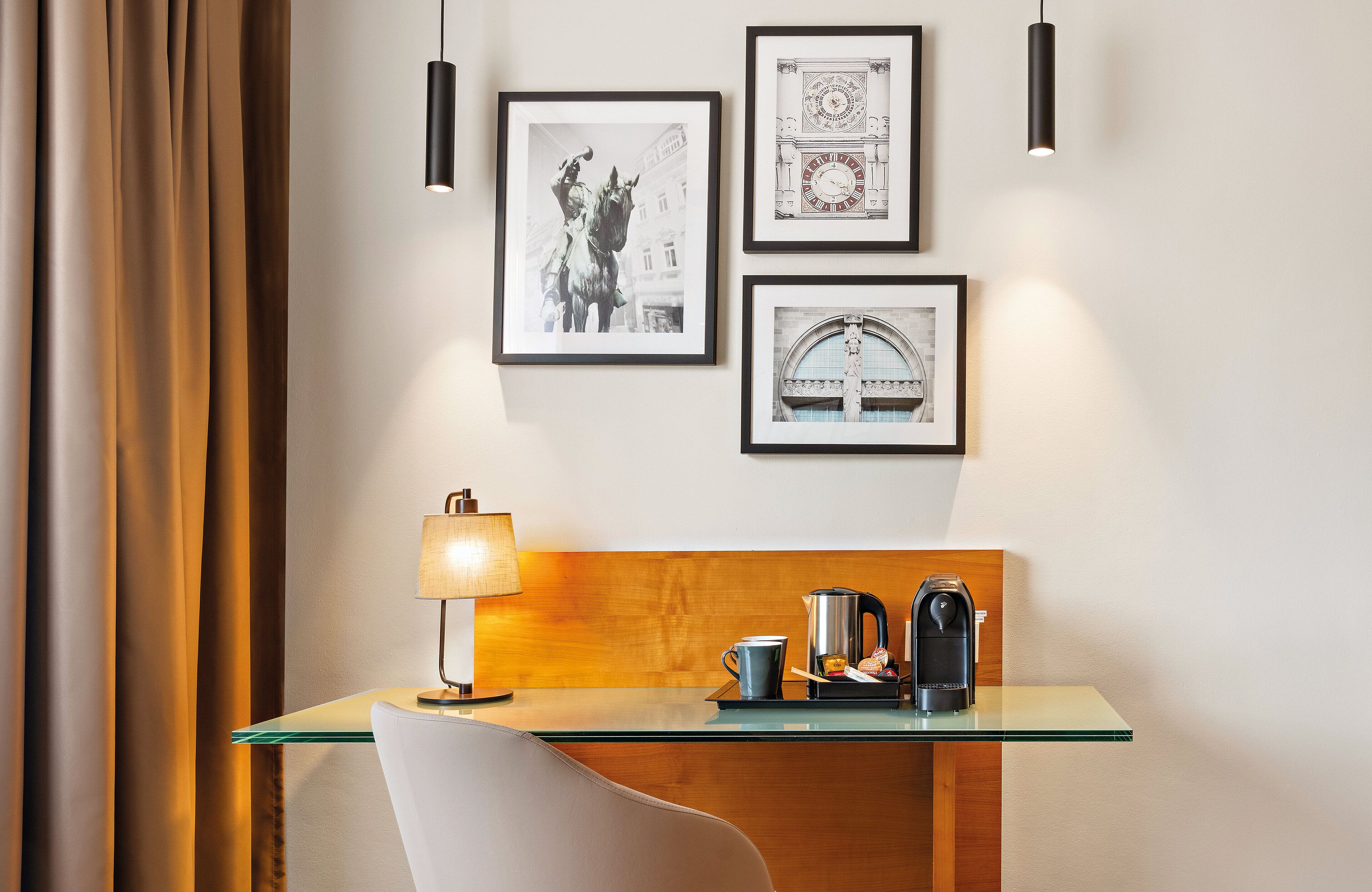 A hotel room with a glass-top desk, a lamp, a coffee maker, and a framed picture of a building.