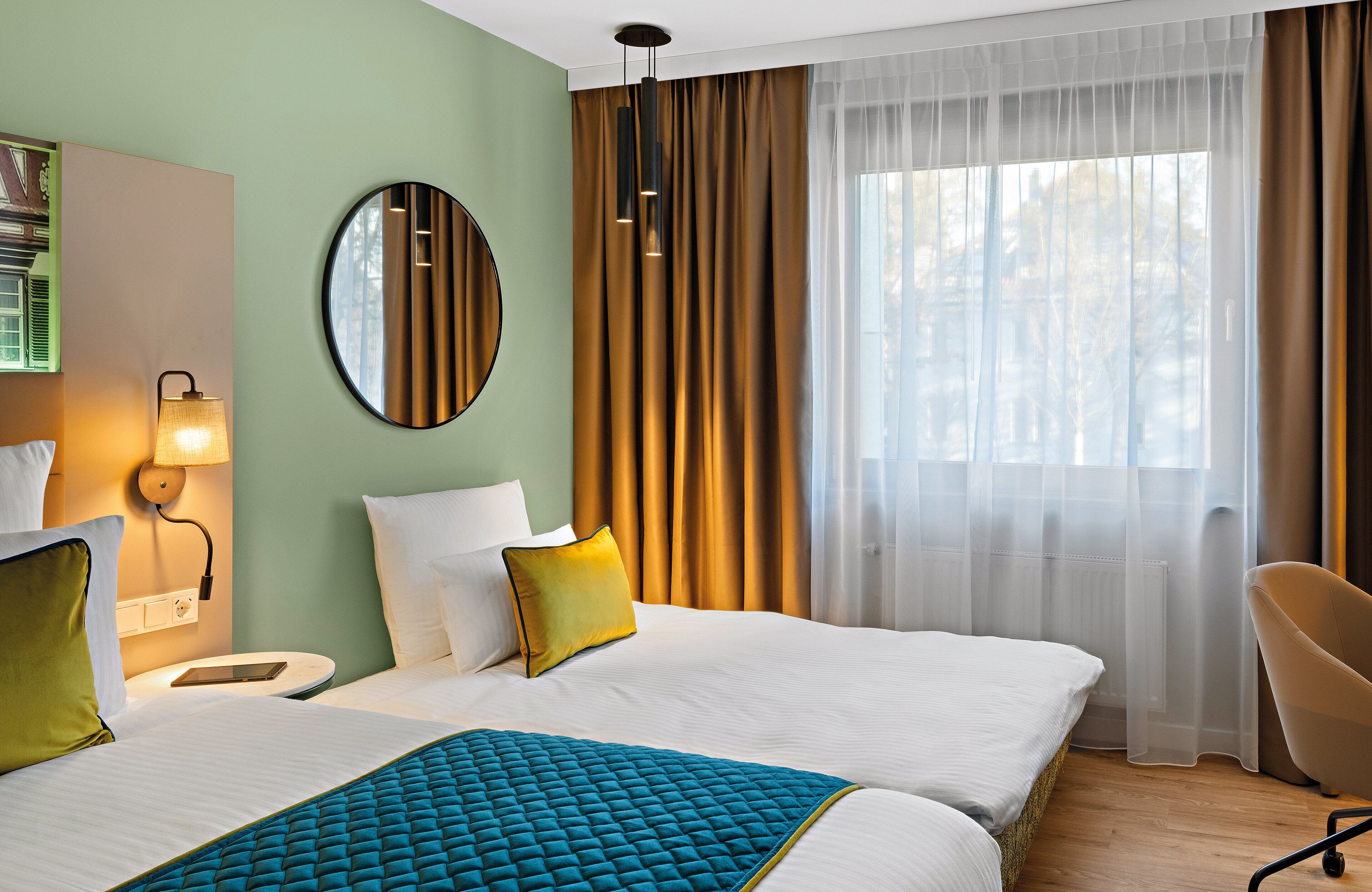 A hotel room with two beds, a round mirror, a bedside table with a lamp, and a chair.