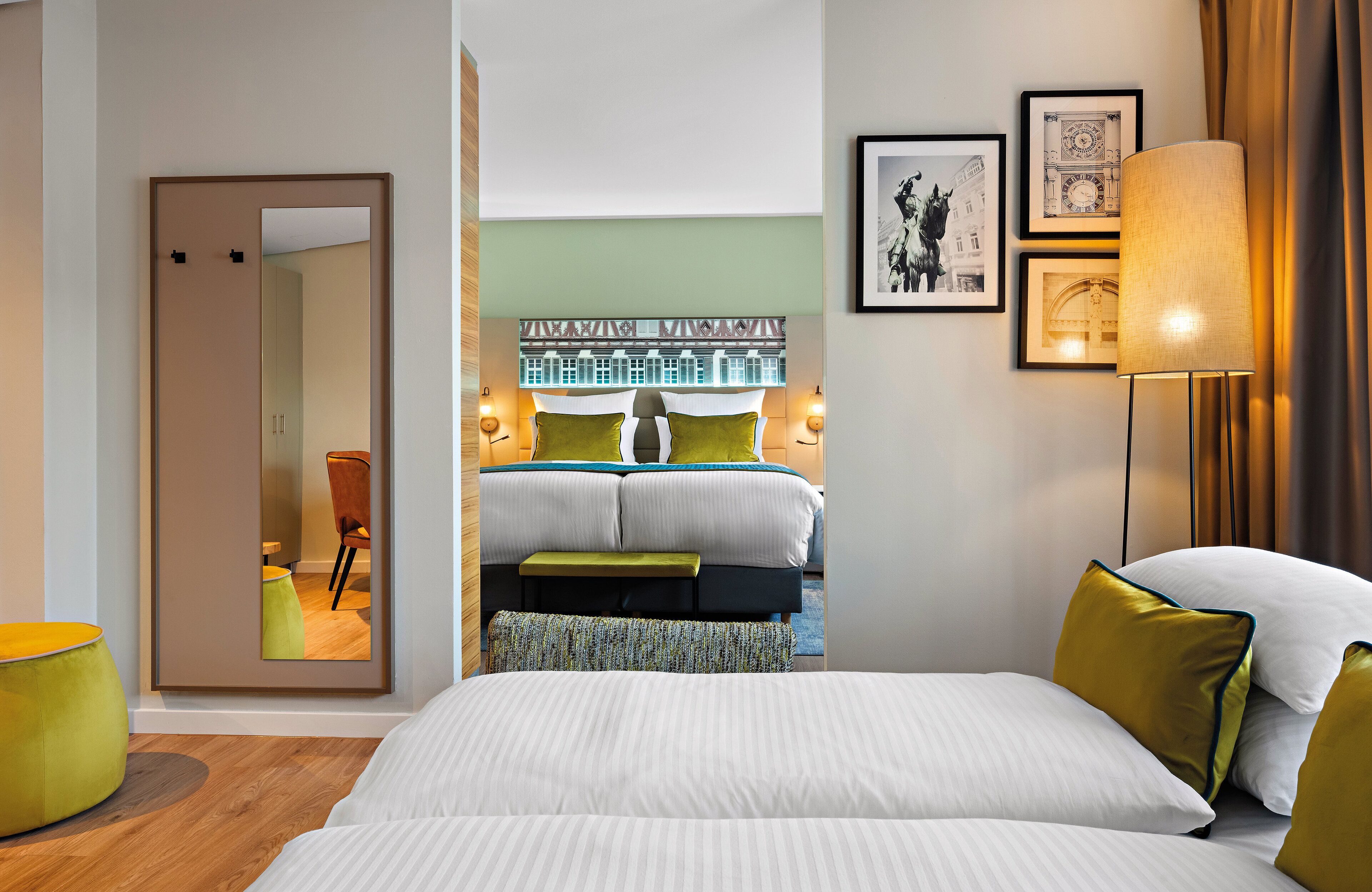 A modern hotel room with a large bed, a mirror, a bench, and a wall-mounted lamp.