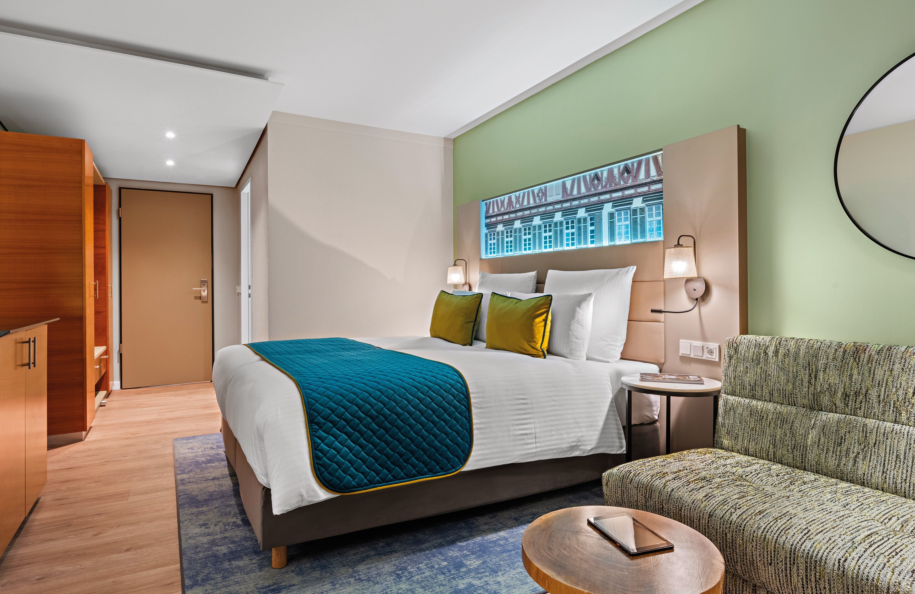 A hotel room with a bed, a nightstand, a chair, and a wall-mounted lamp.