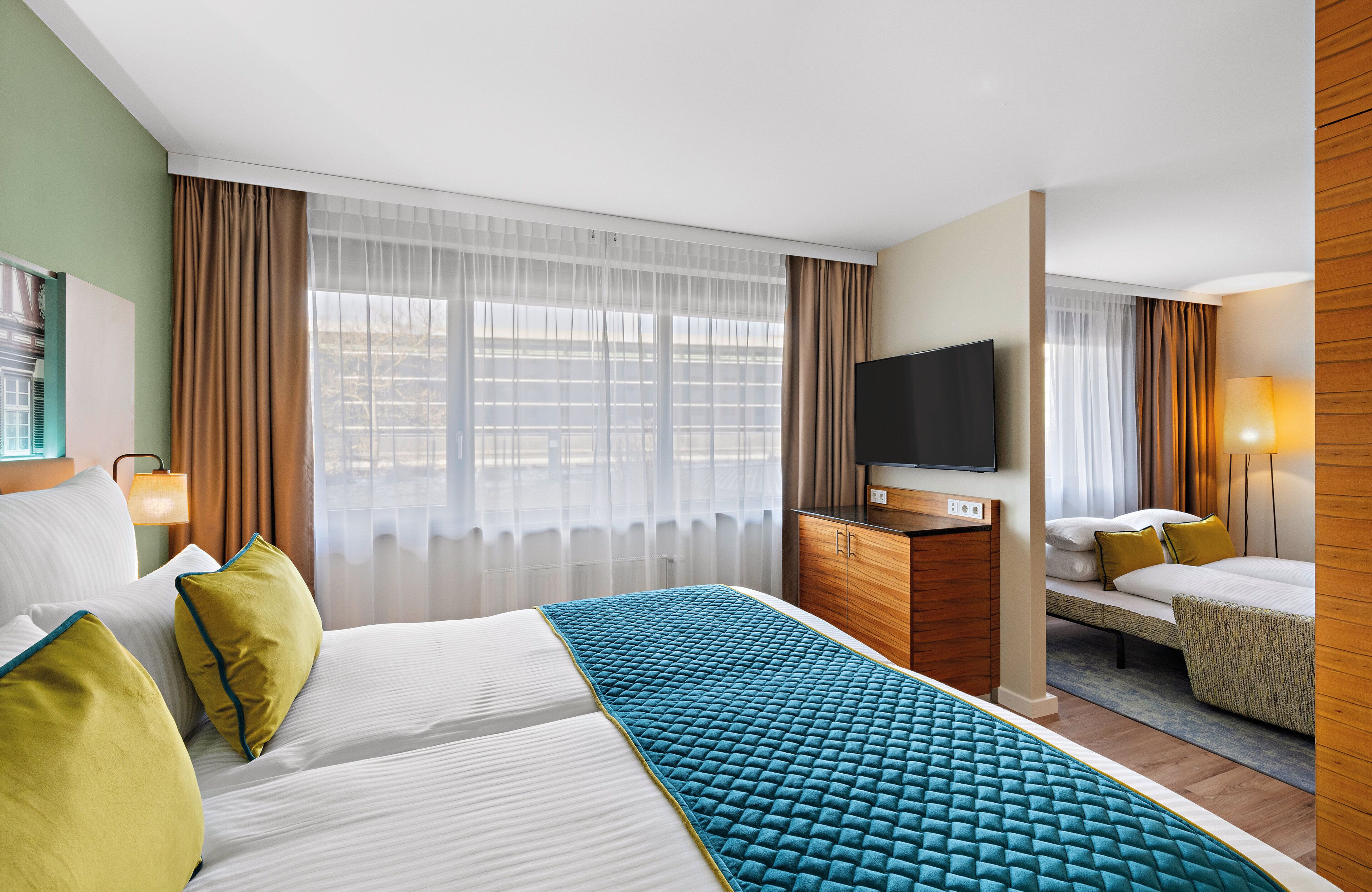 Quarto de hotel com uma cama grande, uma cama menor, uma televisão, uma escrivaninha e uma janela com cortinas.