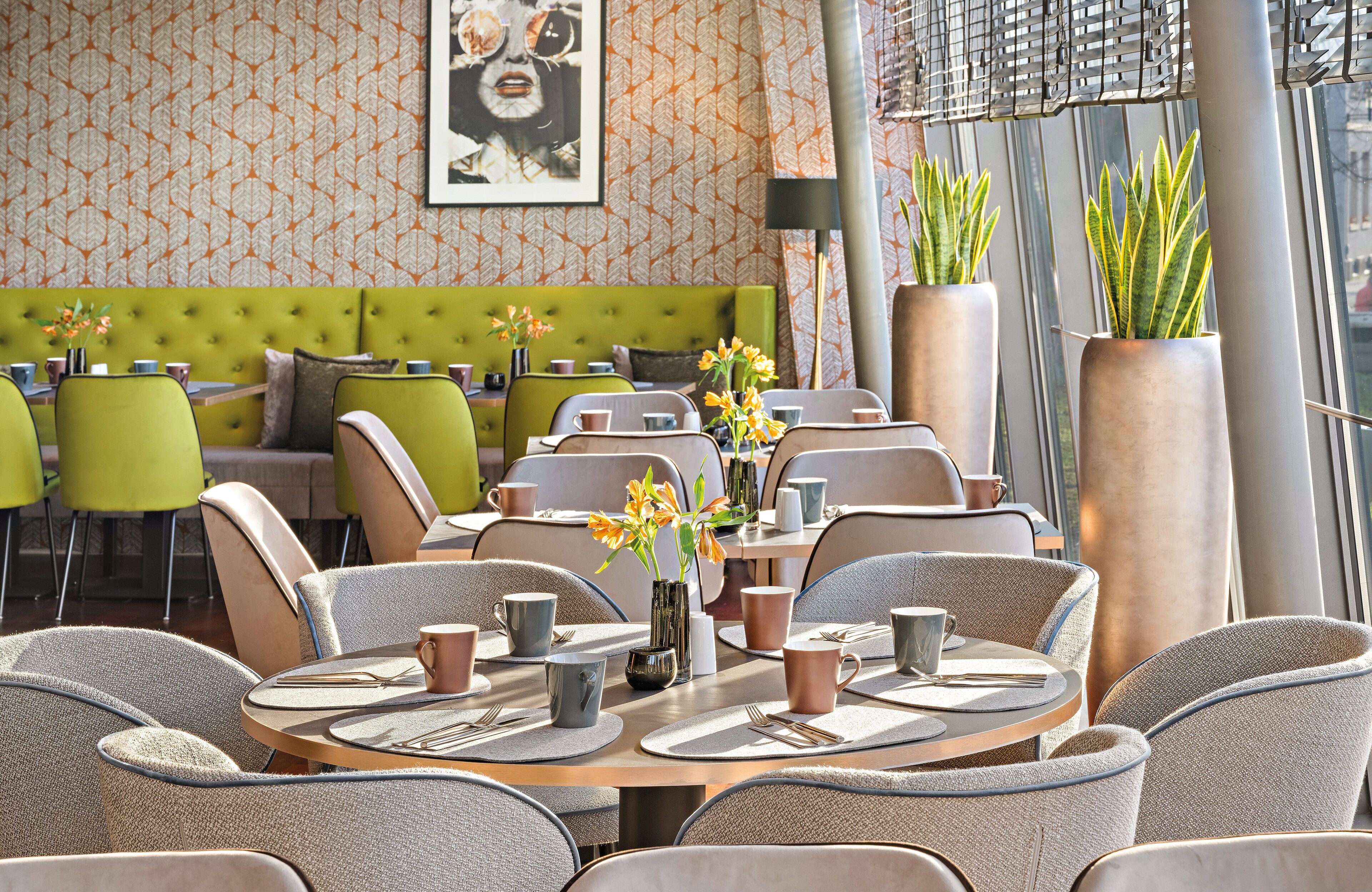 Un restaurant avec des tables rondes, des banquettes recouvertes d’un tissu vert et de grandes plantes en pot.