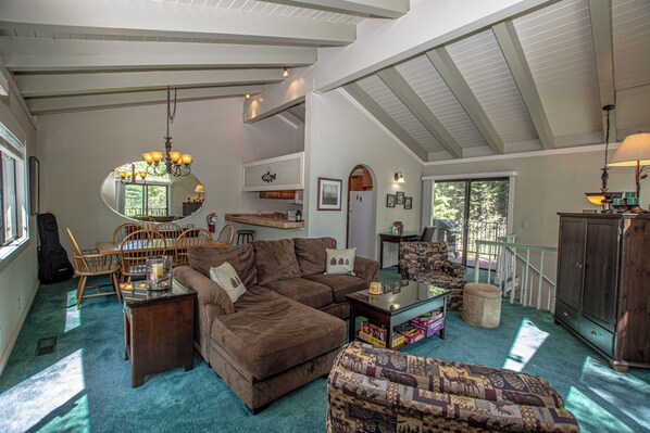 A spacious living room with a brown sectional sofa, a glass coffee table, and a patterned ottoman. There is a dining area with wooden chairs and a small table, and a kitchen area with a wooden cabinet.