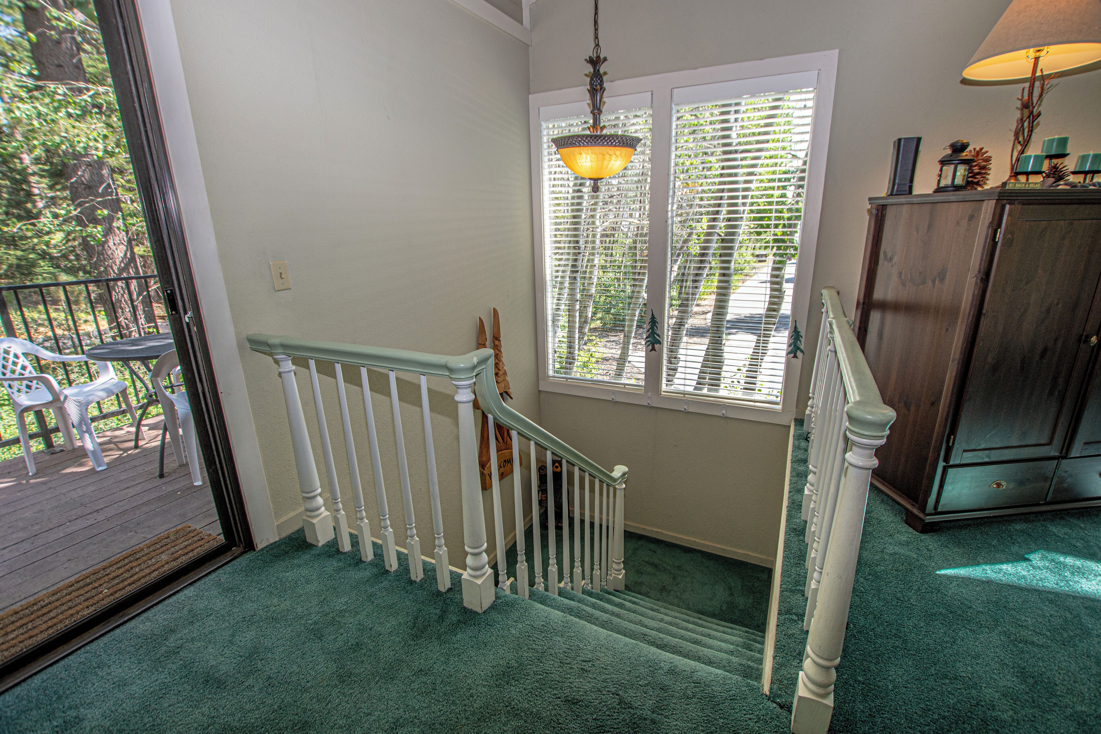Un escalier avec des balustrades blanches, une plateforme recouverte d’une moquette verte et, à travers une porte vitrée, on aperçoit des meubles de jardin.