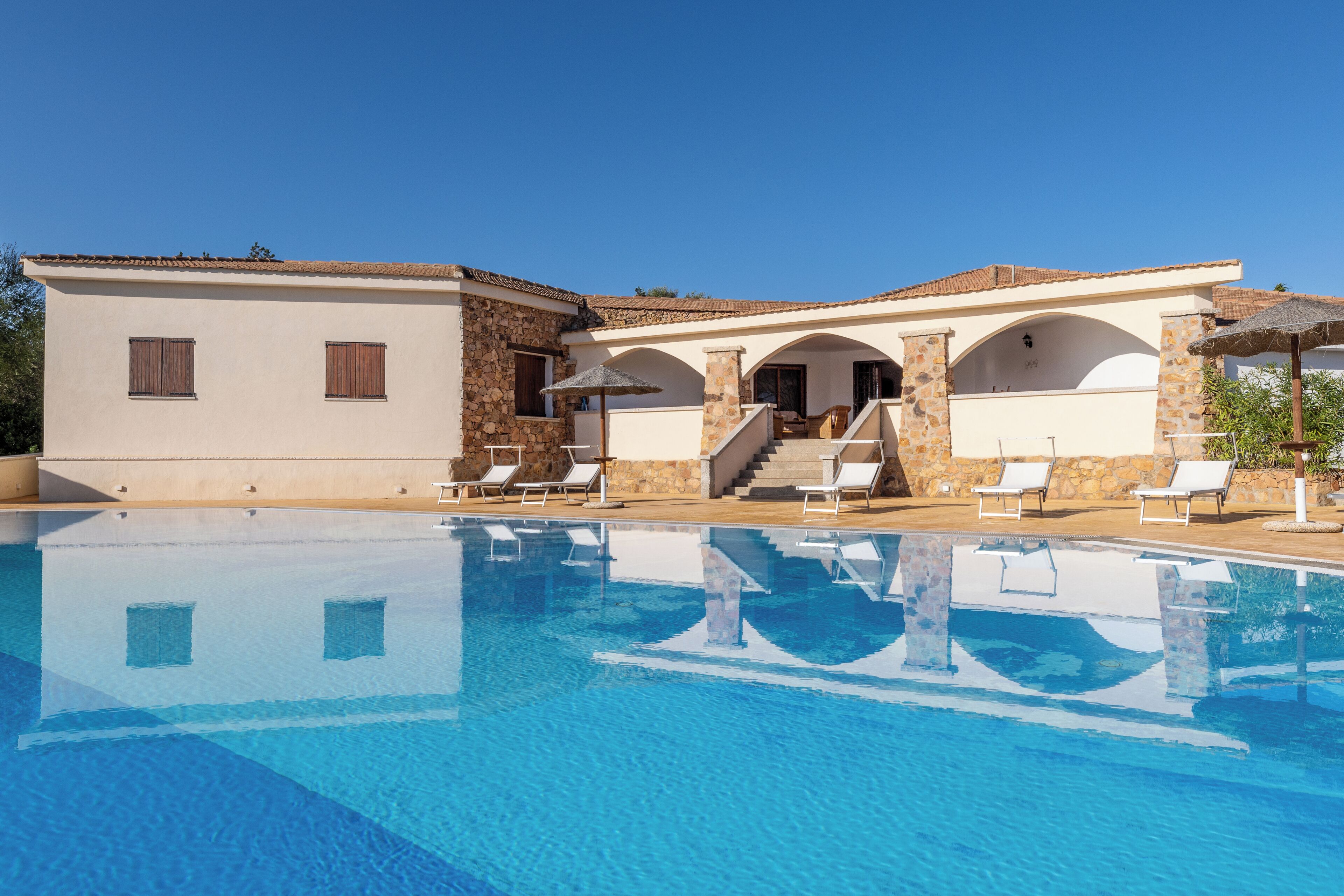 Uma área de piscina com uma piscina azul cristalina, cadeiras de sol brancas e um edifício de pedra com arcos e telhado em azulejos.