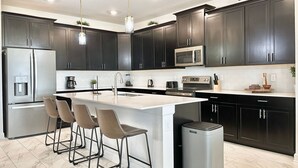 Una cocina moderna con armarios oscuros, electrodomésticos de acero inoxidable, una isla central con taburetes altos y un cubo de basura.