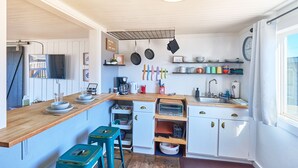 Une cuisine compacte avec un plan de travail en bois, un évier et un petit réfrigérateur. On y trouve des tabourets et une étagère avec des ustensiles de cuisine.