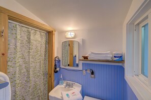 Une salle de bain avec une porte en bois, un rideau de douche orné de motifs de feuilles, un lavabo avec un miroir au-dessus, et une étagère avec des serviettes et un distributeur de savon.