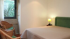 Ein Schlafzimmer mit Bett, einem Sessel, einem Nachttisch mit Lampe und einem Fenster mit Blick auf Bäume.