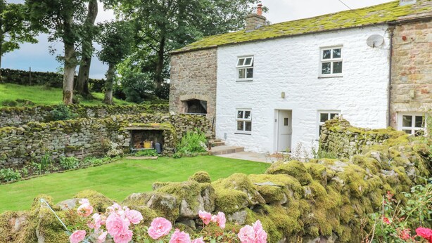 A stone house with a white exterior, a stone wall with a moss-covered rock, and pink flowers in the foreground. - The La'al Cragg (Kirkby Stephen)