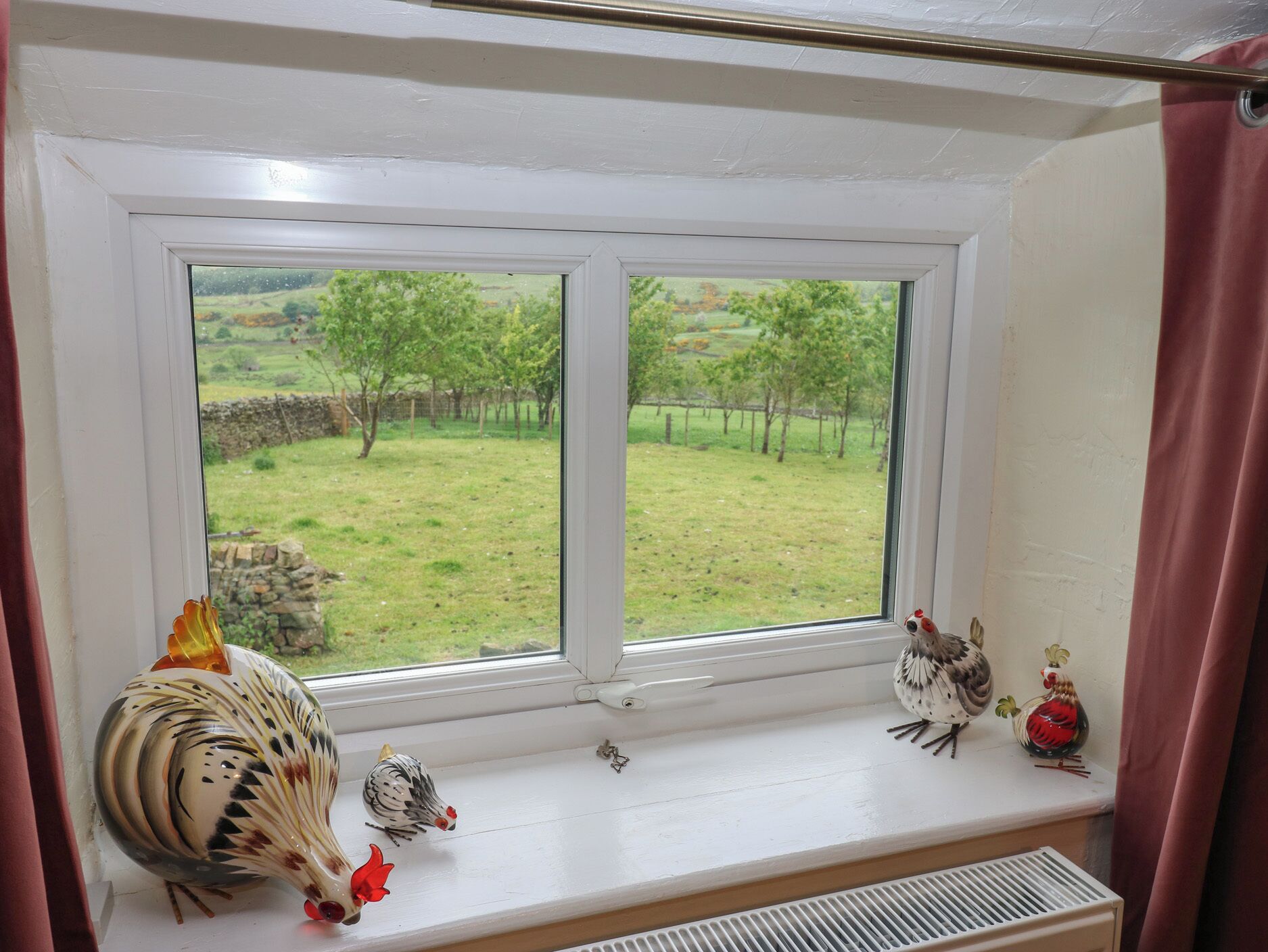 Una ventana con vistas a un campo cubierto de hierba y árboles, una rejilla de ventilación debajo, y gallinas decorativas en el alféizar.
