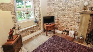 A stone-walled room with a television on a wooden stand, a window with a view of greenery, and a fireplace. - The La'al Cragg (Kirkby Stephen)