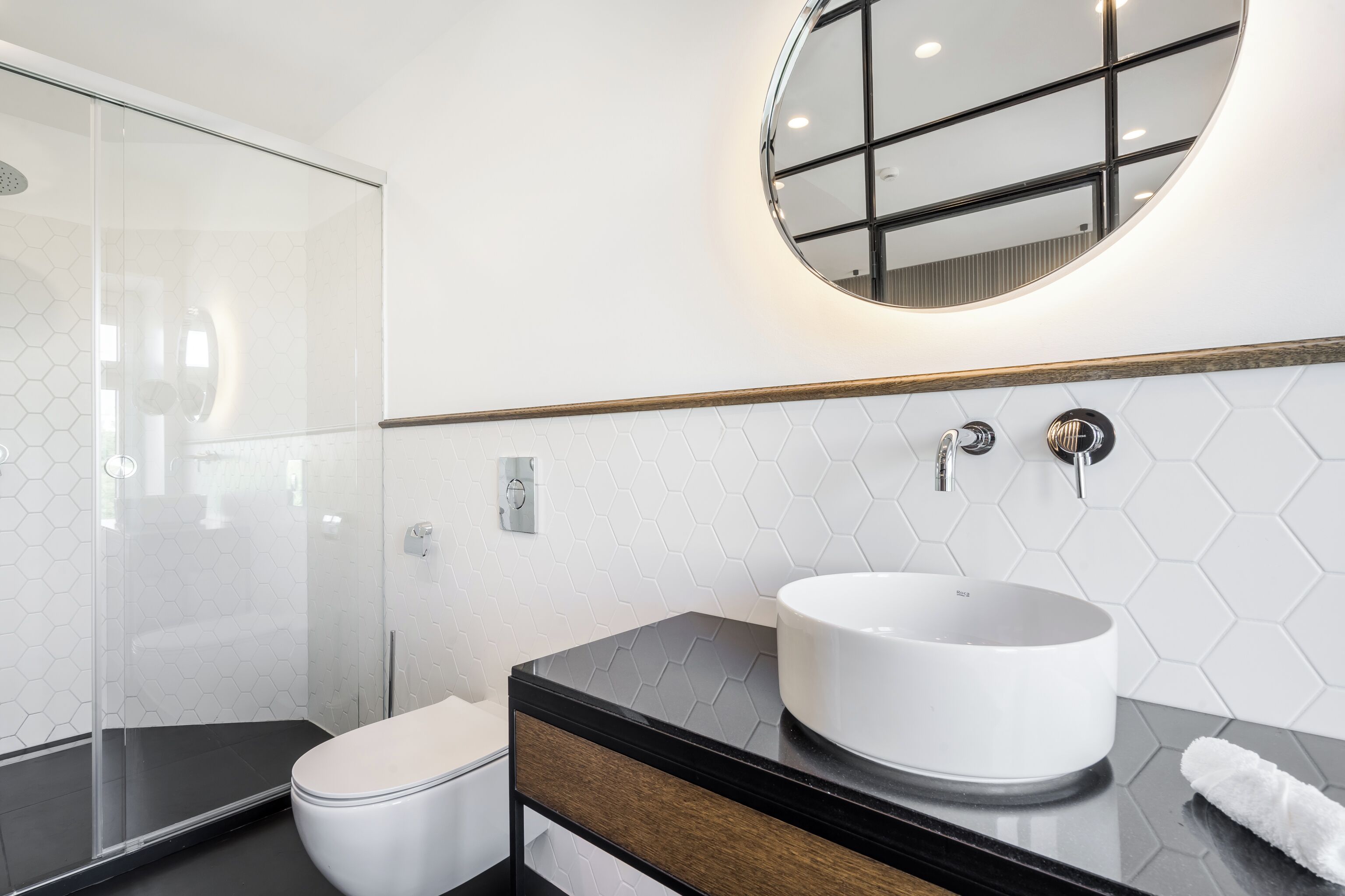 A modern bathroom with a glass shower enclosure, a white toilet, a black vanity with a round mirror, and a white sink.