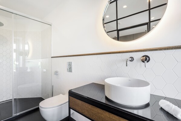A modern bathroom with a glass shower enclosure, a white toilet, a black vanity with a round mirror, and a white sink.