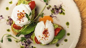 Een bord met een gerecht bestaande uit avocado plakjes, tomaten en een gepocheerd ei, versierd met microgroenten en een beetje groene saus.
