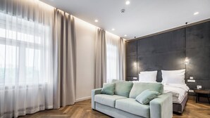 Een moderne slaapkamer met een lichtkleurige bank, een bed met witte beddengoed en een groot raam met dunne gordijnen.