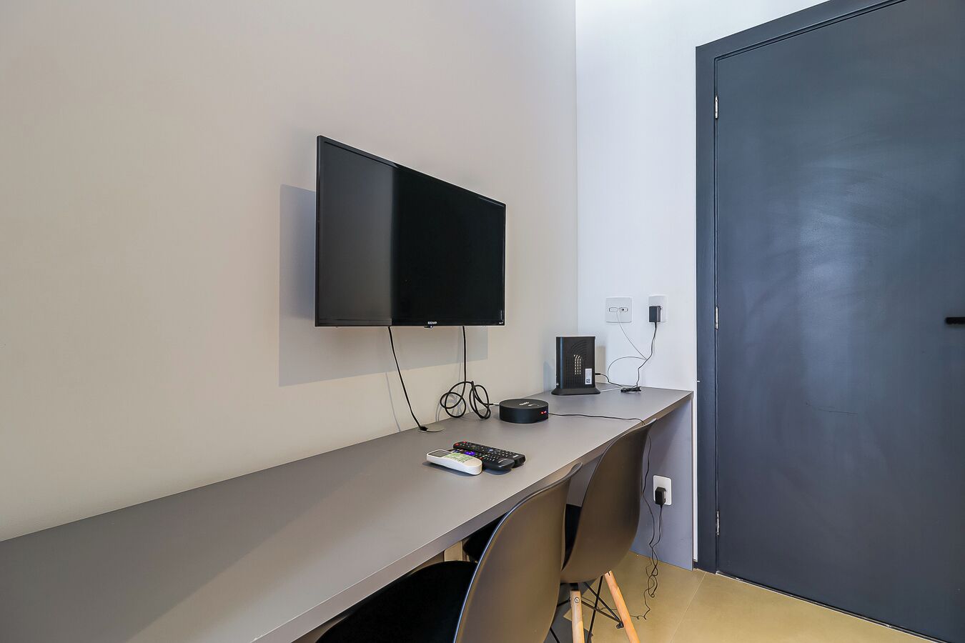 Una habitación de hotel moderna con un televisor de pantalla plana montado en la pared, un escritorio con una computadora y un control remoto, y una puerta con manija.